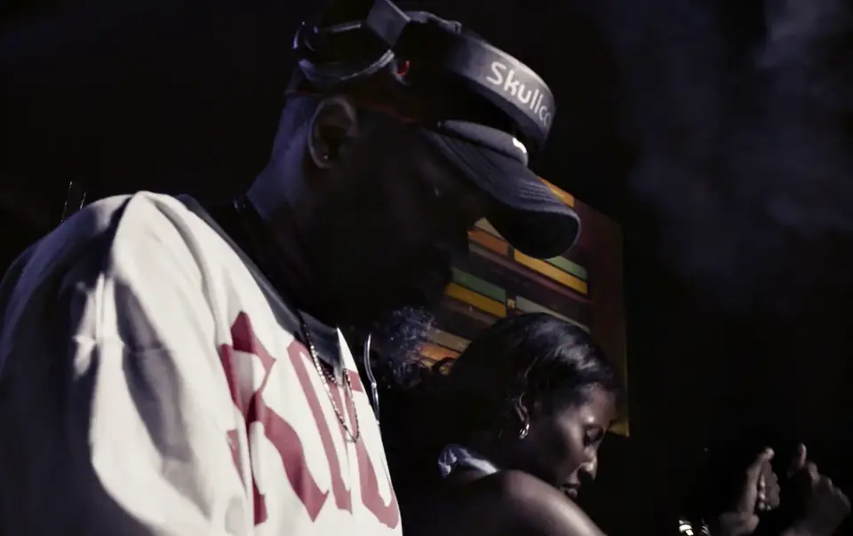 A DJ in a cap and a white shirt at the decks, with a woman dancing close behind him